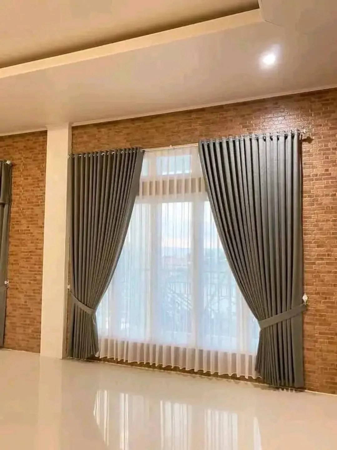 Gorden abu-abu dengan vitrase putih pada jendela di ruangan berdinding motif bata